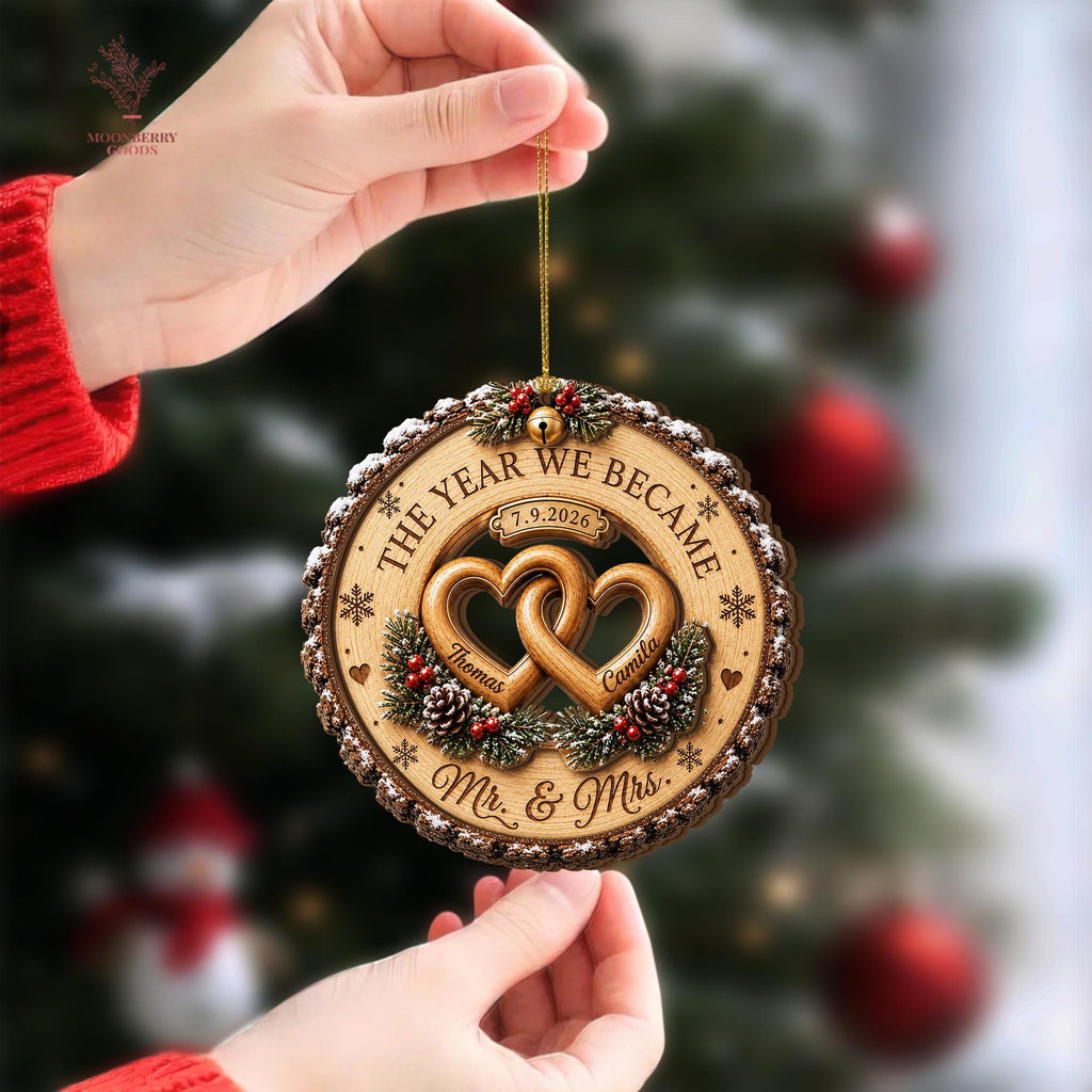 May include: A round wooden Christmas ornament featuring two interlocking hearts in the center, engraved with the names Thomas and Camila, the date 7.9.2026, and the text THE YEAR WE BECAME Mr. & Mrs. surrounded by pine cones and red berries.