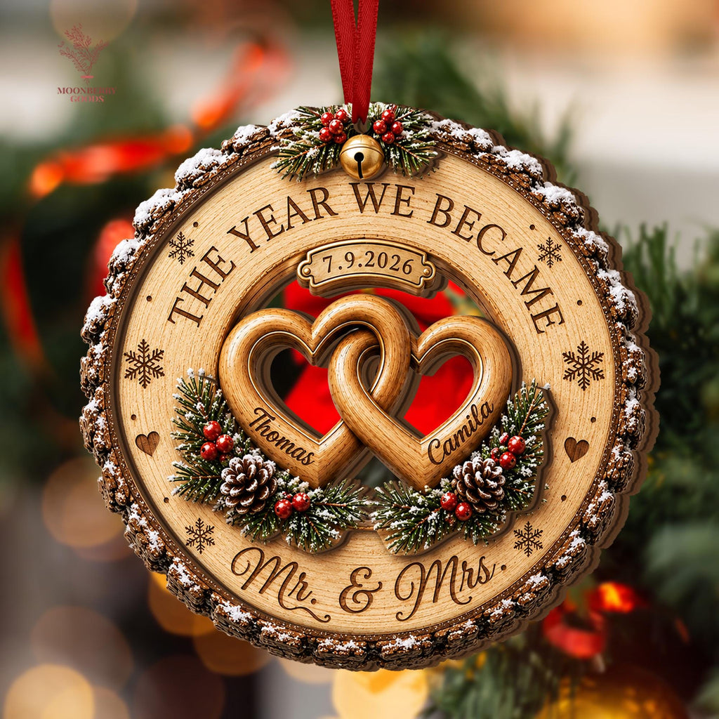 May include: A round wooden Christmas ornament featuring two interlocking hearts engraved with the names Thomas and Camila. The text reads THE YEAR WE BECAME, 7.9.2026, and Mr. & Mrs. It is decorated with pine branches, red berries, pinecones, and a gold bell.
