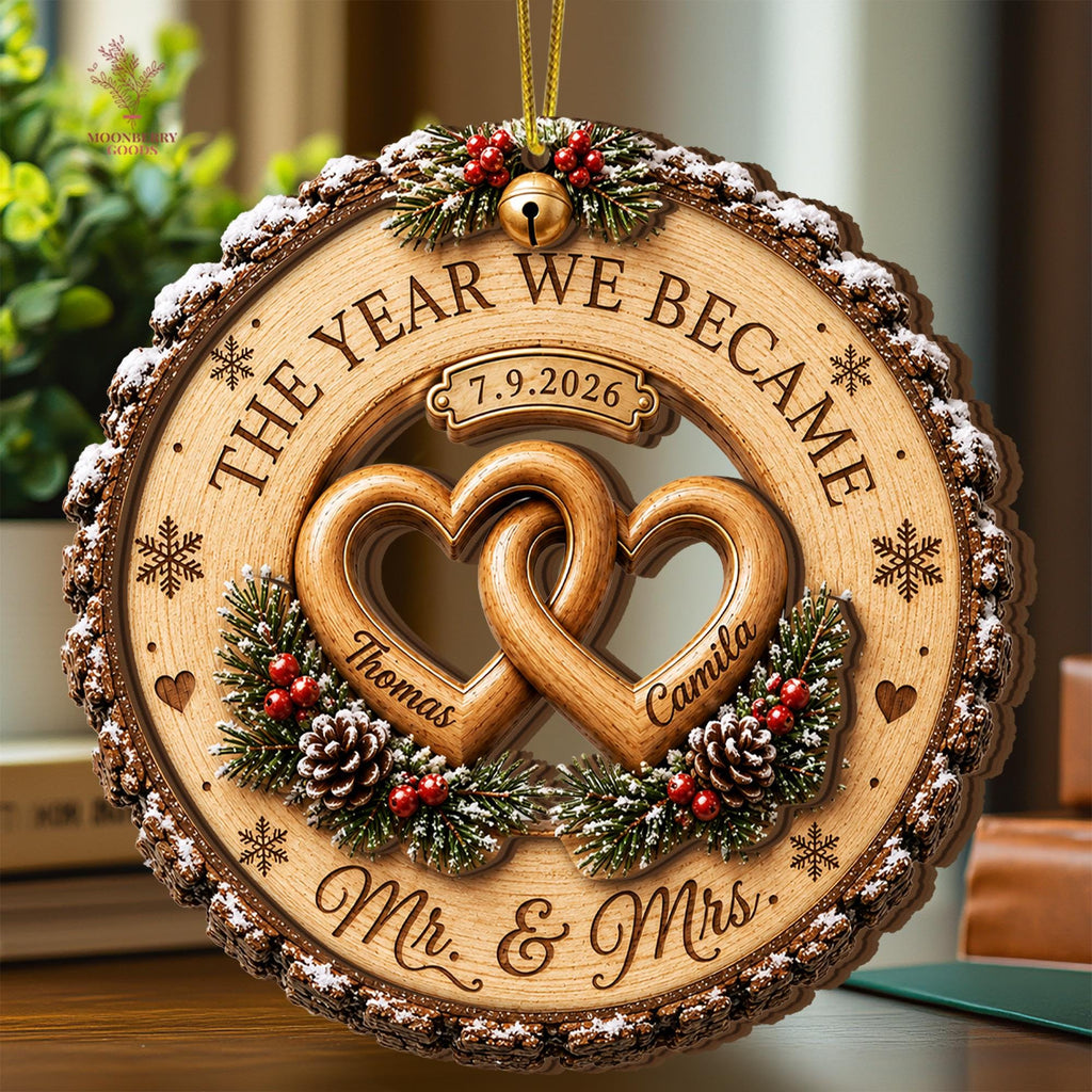 May include: A circular wooden ornament featuring two interlocking hearts engraved with the names Thomas and Camila. The text reads THE YEAR WE BECAME, 7.9.2026, and Mr. & Mrs. It is decorated with pine branches, red berries, pinecones, and a gold bell.