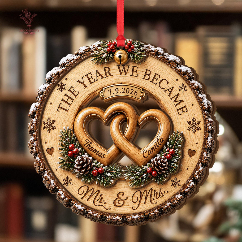 May include: A round wooden ornament with bark edges featuring two interlocking hearts engraved with the names Thomas and Camila. The text reads THE YEAR WE BECAME, 7.9.2026, and Mr. & Mrs. It is decorated with pine branches, red berries, and a gold bell.