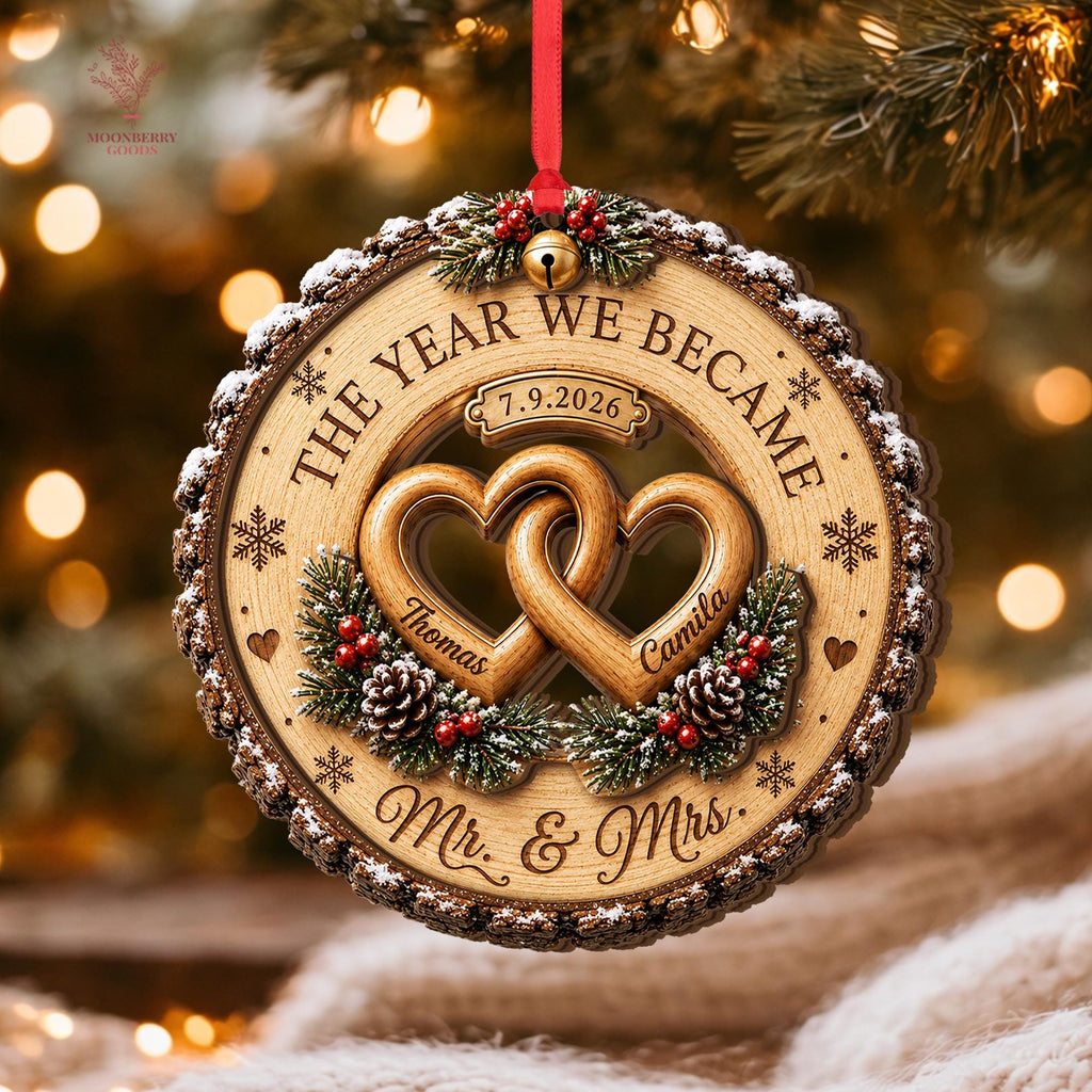 May include: A round wooden Christmas ornament with bark edges, featuring two interlocking heart cutouts engraved with the names Thomas and Camila. The text reads THE YEAR WE BECAME, 7.9.2026, and Mr. & Mrs., accented by pine branches, red berries, and snowflakes.