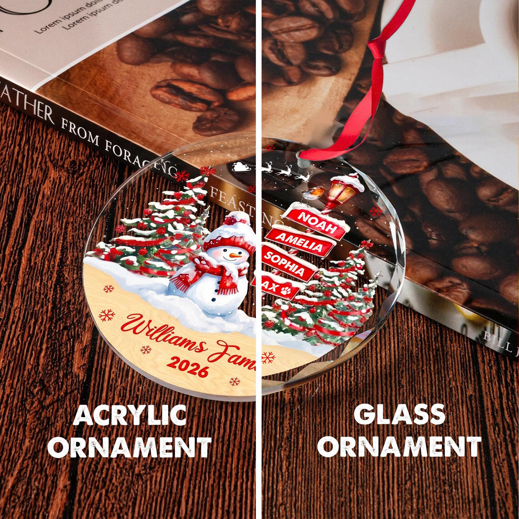 May include: A round, clear ornament featuring a festive winter scene with a snowman, snow-covered pine trees, and red accents. The text reads Williams Family 2026, with additional name signs for Noah, Amelia, Sophia, and Max. It includes a red ribbon.
