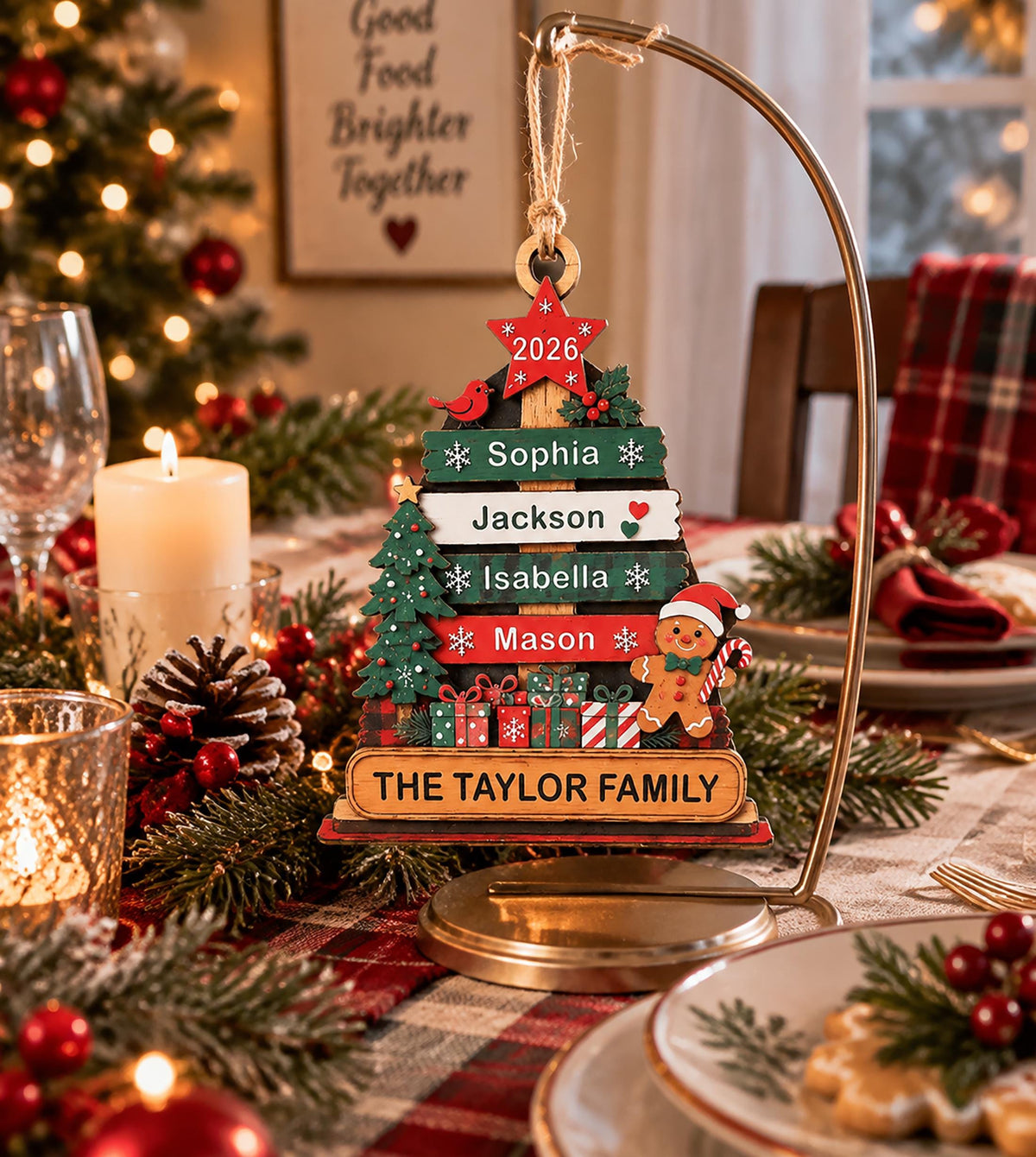 May include: A wooden Christmas tree ornament hanging from a gold metal stand. The tree features horizontal slats with the names Sophia, Jackson, Isabella, and Mason, a red star labeled 2026, a gingerbread man, and a base reading THE TAYLOR FAMILY.