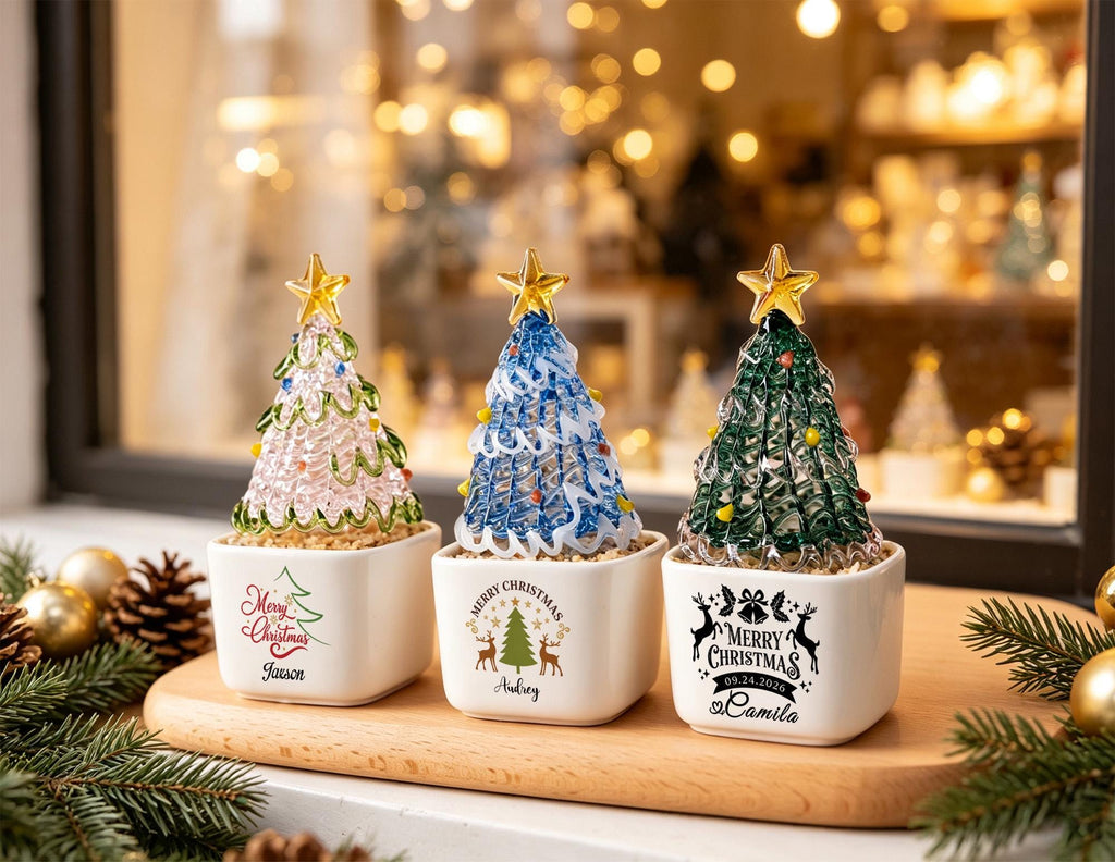 May include: Three glass Christmas tree ornaments in white square ceramic pots. The trees are pink, blue, and green with gold stars on top. Each pot features custom text and holiday graphics including Merry Christmas, names, and the date 09.24.2026.
