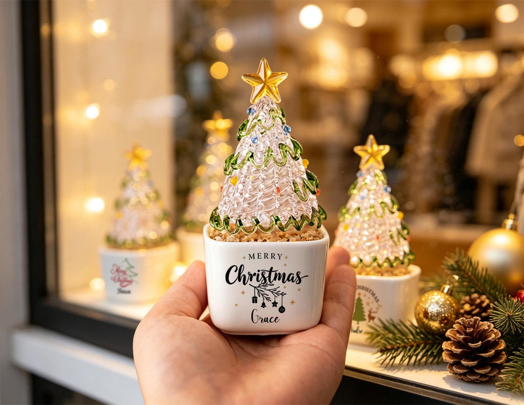 May include: A clear glass Christmas tree figurine with green glass garland and a gold star topper, set in a white square ceramic planter. The planter features the black text Merry Christmas and the name Grace, accented with small black ornaments.