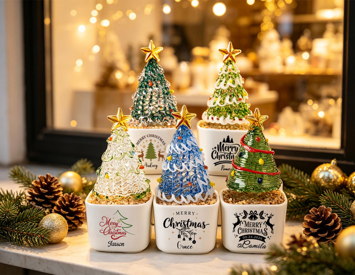 May include: Five decorative glass Christmas trees in white square ceramic pots. Each tree features a gold star topper and colorful glass accents. The pots are printed with various holiday designs, including Merry Christmas text and names.
