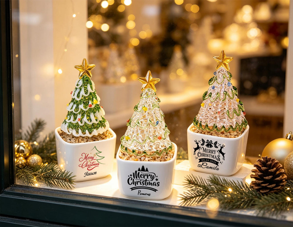 May include: Three glass Christmas tree figurines in white square ceramic pots. Each tree is topped with a gold star and decorated with green swirls and colorful dots. The pots feature black or red text including Merry Christmas and names.