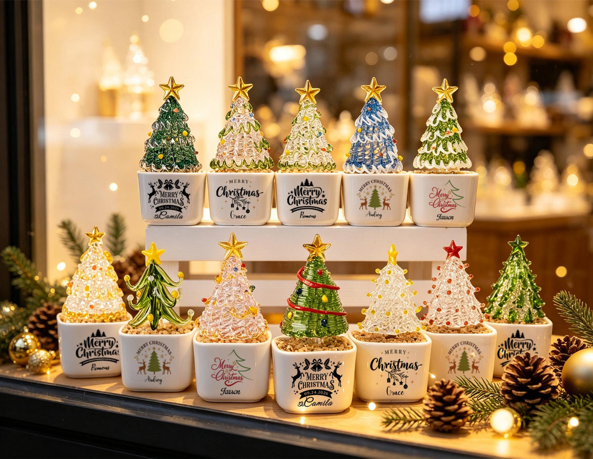 May include: A collection of eleven miniature glass Christmas tree figurines, each mounted on a square white ceramic base. The trees feature various colors including green, clear, blue, and pink, topped with gold stars. Each base displays Merry Christmas text.
