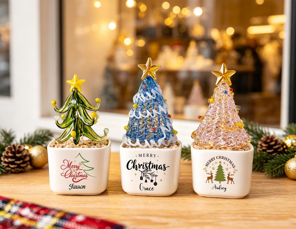 May include: Three glass Christmas tree figurines in white square ceramic pots. The left pot says Merry Christmas and Jaxson, the center says Merry Christmas and Grace, and the right says Merry Christmas and Audrey. The trees are green, blue, and pink.