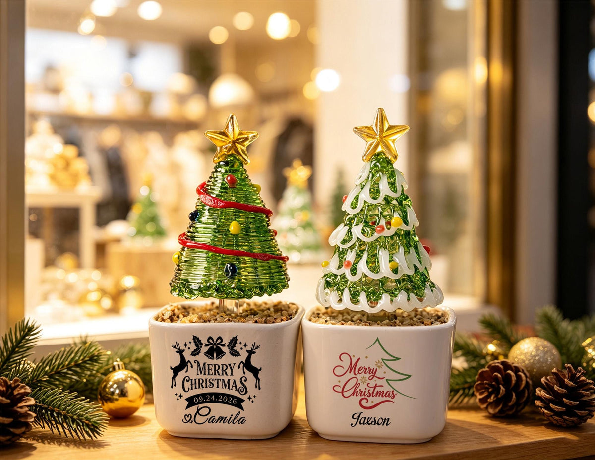 May include: Two glass Christmas tree ornaments in white square ceramic pots. The left pot features black text reading Merry Christmas 09.24.2026 Camila with reindeer. The right pot features red and green text reading Merry Christmas and the name Jaxson.