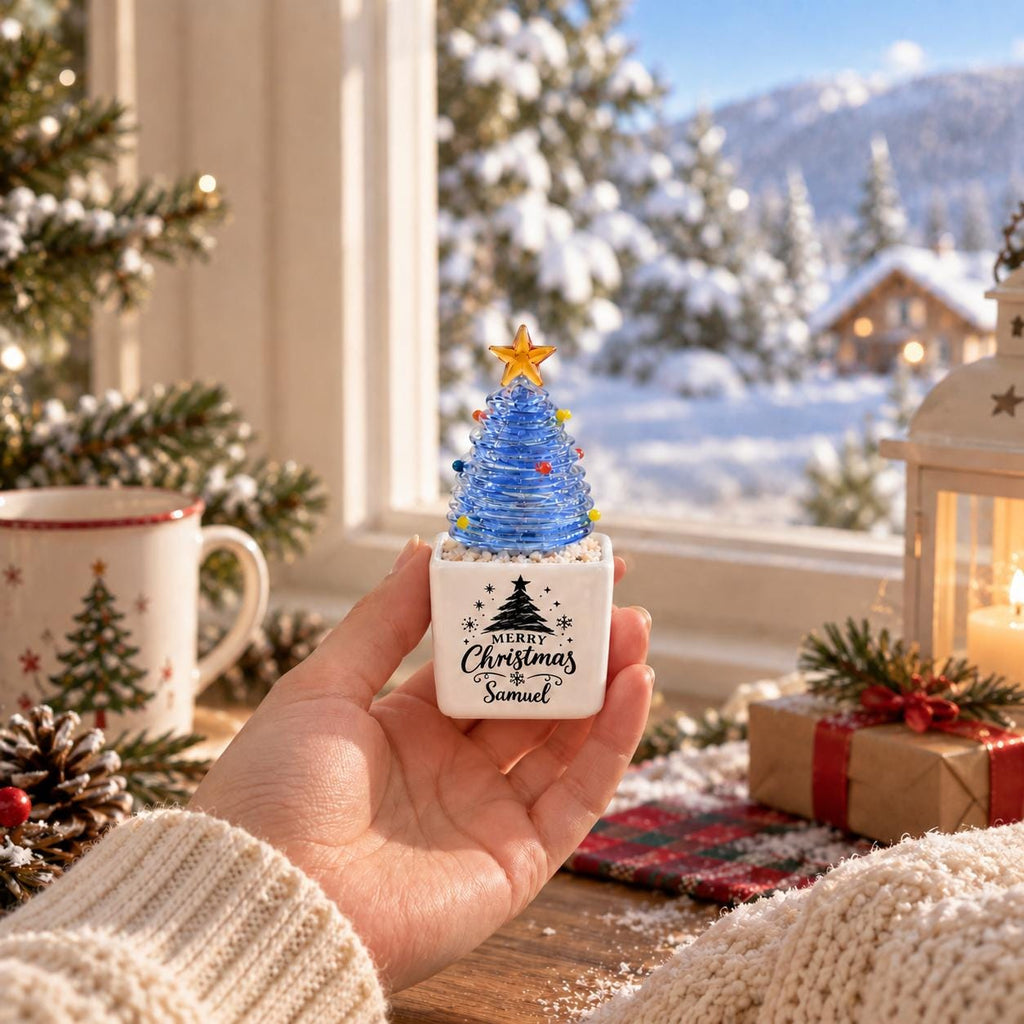 May include: A small blue glass Christmas tree ornament with a yellow star topper, set in a white square ceramic base. The base features a black printed graphic of a pine tree and the text Merry Christmas Samuel in a decorative script font.