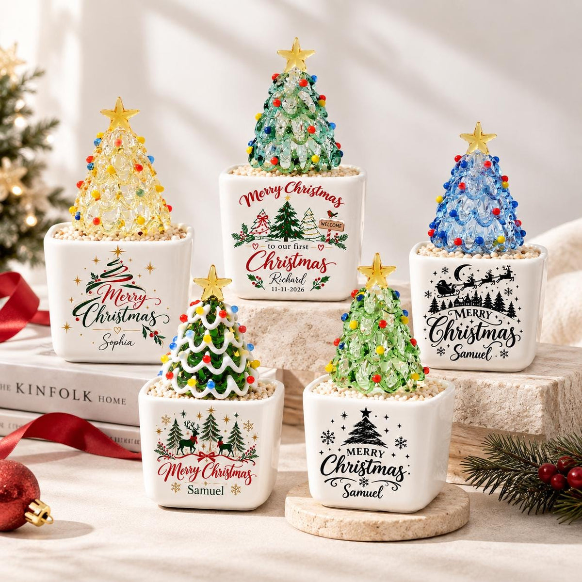 May include: Five square white ceramic planters, each containing a small glass Christmas tree with a yellow star topper and colorful lights. The planters feature various black and red holiday graphics and text, including Merry Christmas and names.