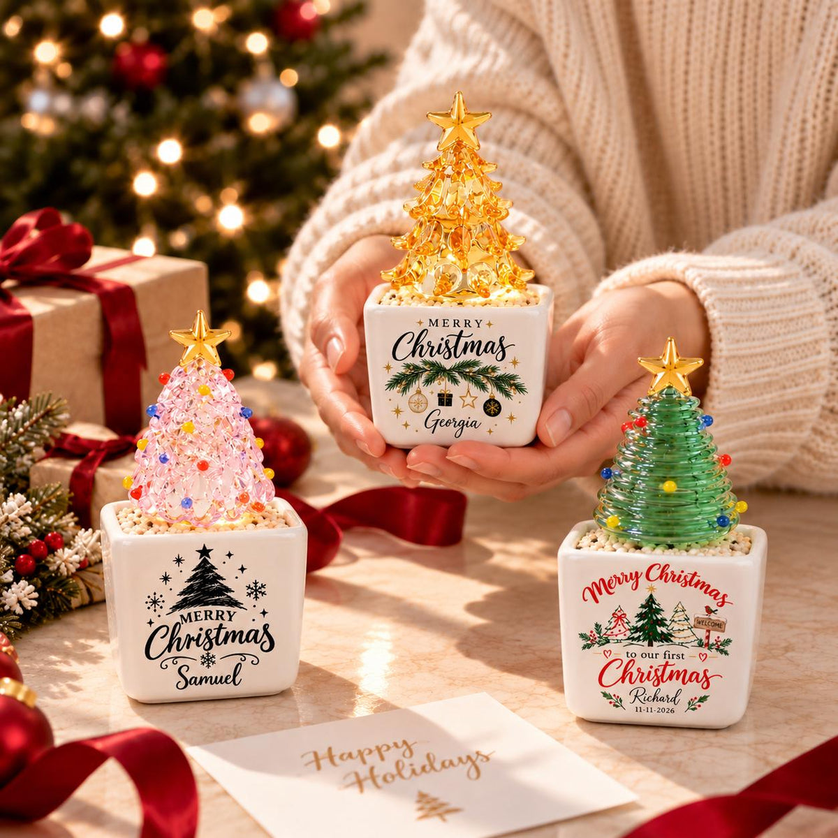 May include: Three decorative tabletop Christmas trees in square white ceramic pots. One tree is amber, one is pink, and one is green, each topped with a gold star. The pots feature festive text and graphics, including the names Georgia, Samuel, and Richard.
