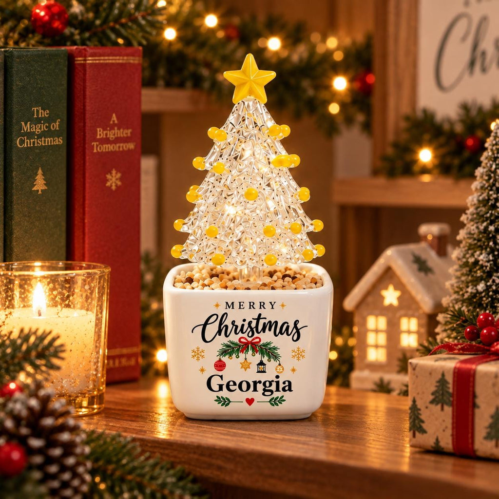 May include: A clear, textured glass Christmas tree ornament with yellow spherical accents and a yellow star topper, set in a white square ceramic planter. The planter features the text Merry Christmas Georgia with festive holiday graphics.