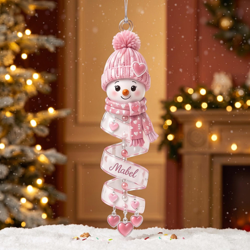 May include: A hanging Christmas ornament featuring a snowman head wearing a pink knit hat with a pom-pom and a matching scarf. A spiral ribbon hangs below, decorated with small pink hearts and the name Mabel, ending in three dangling heart charms.