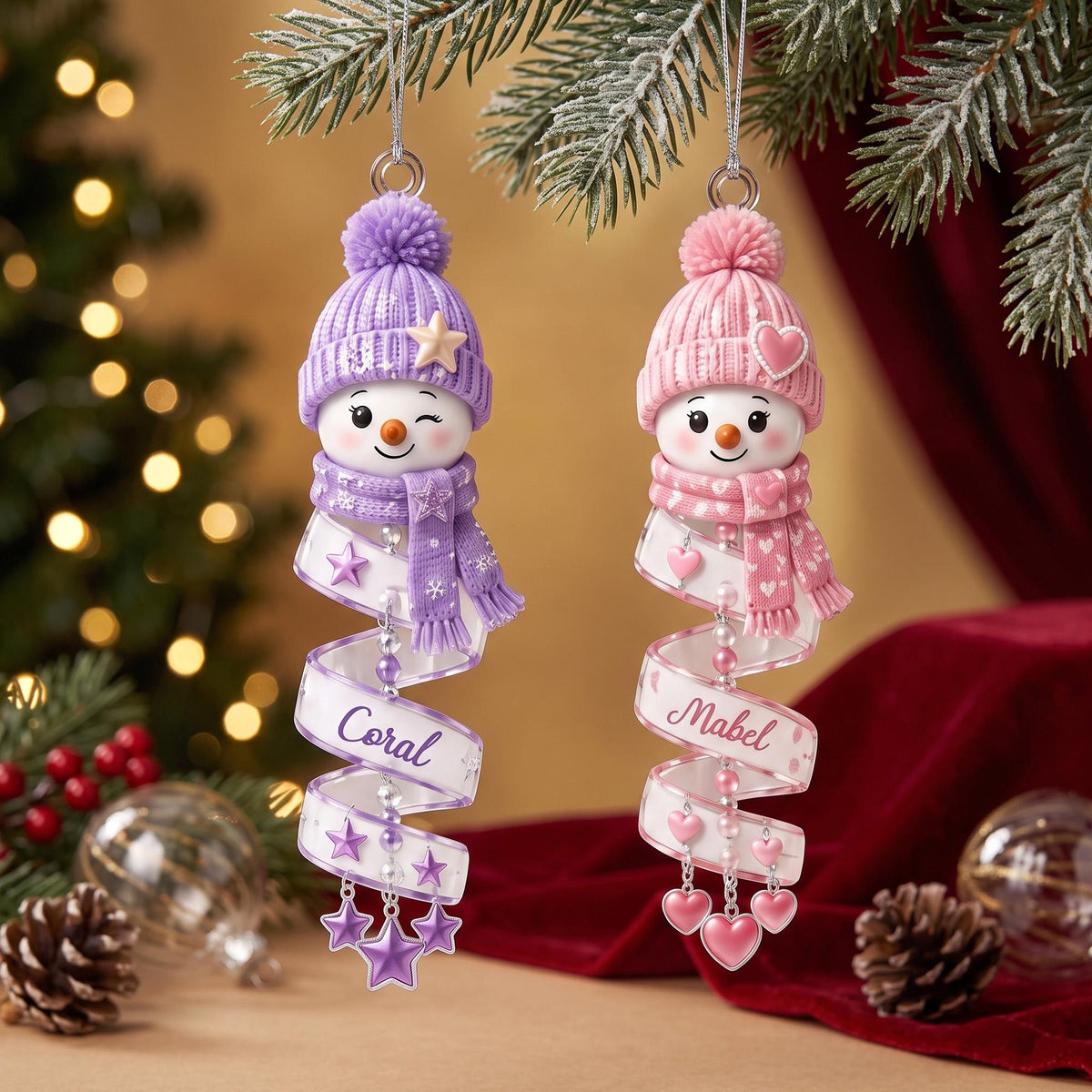 May include: Two hanging snowman ornaments with pom-pom hats and scarves. The left snowman wears a purple hat and scarf with a star, and a ribbon labeled Coral. The right snowman wears a pink hat and scarf with a heart, and a ribbon labeled Mabel.