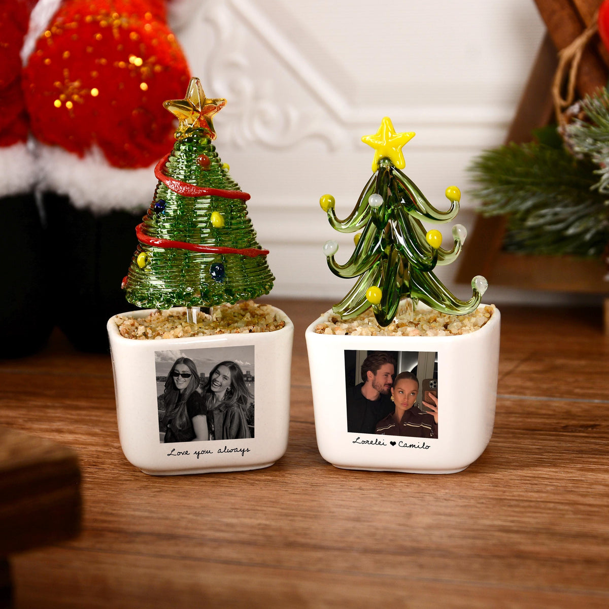 May include: Two white square ceramic planters containing glass Christmas trees. The left tree is green with a red garland and yellow star. The right tree is green with yellow ornaments and a yellow star. Each planter features a black and white photo.