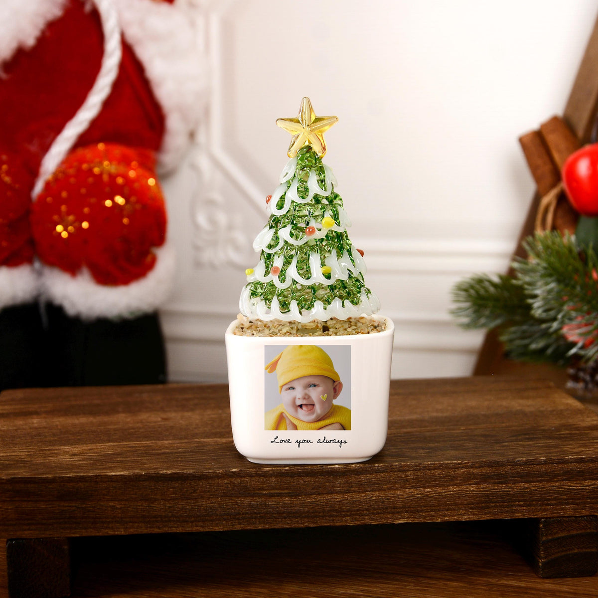 May include: A white square ceramic planter featuring a photograph of a smiling baby wearing a yellow hat, with the text "Love you always" printed below. A glass Christmas tree with green textured foliage and a gold star topper sits inside.