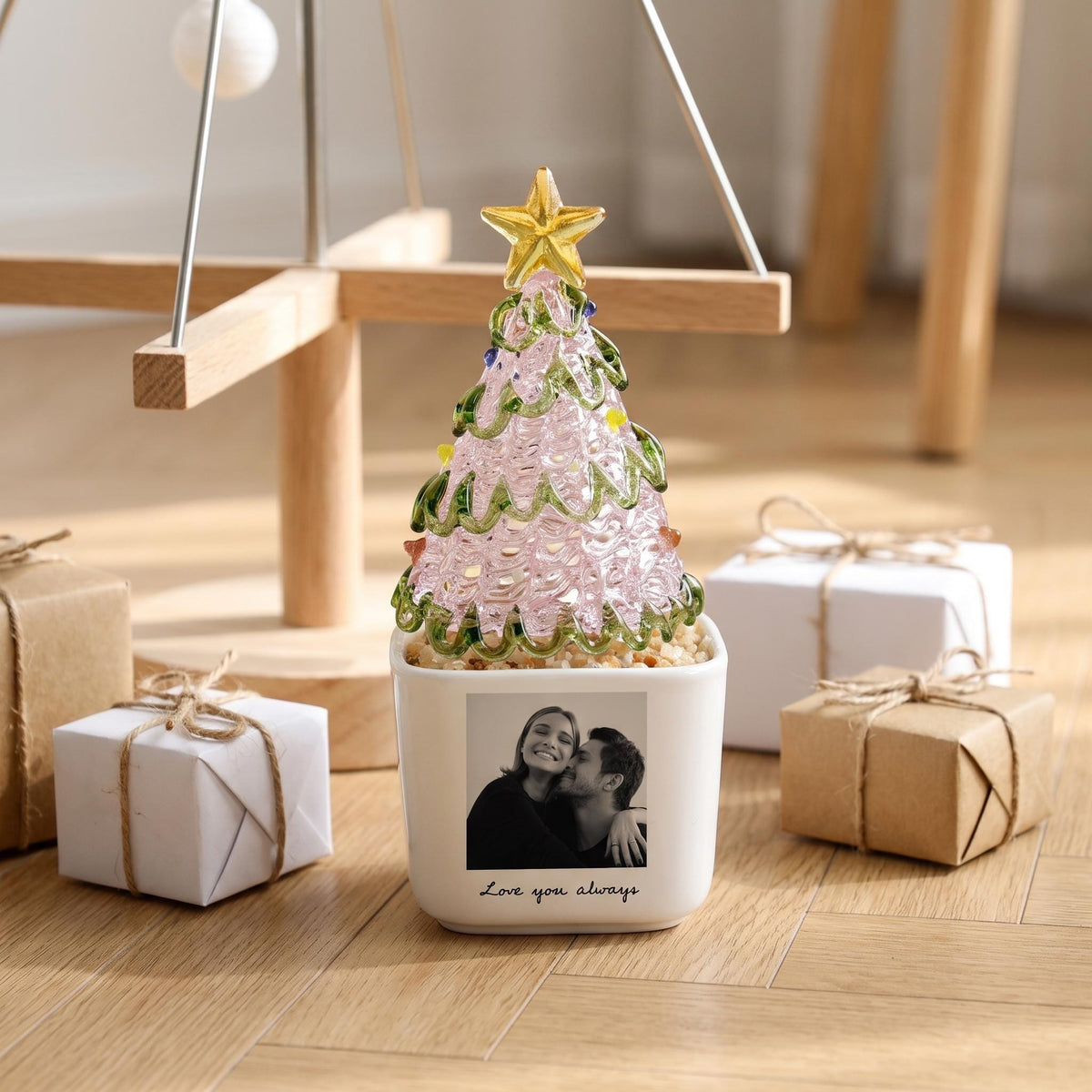 May include: A decorative glass Christmas tree ornament with a pink textured body, green glass branches, and a gold star topper. The tree sits in a white square ceramic planter featuring a black and white photograph of a couple and the text "Love you always."