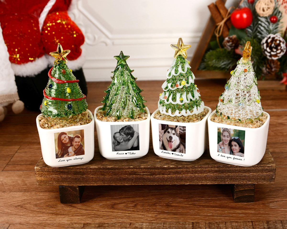 May include: Four glass Christmas tree ornaments in white square ceramic pots. Each tree features a gold star topper and sits on a bed of small pebbles. The pots are personalized with black and white photographs and text captions at the base.