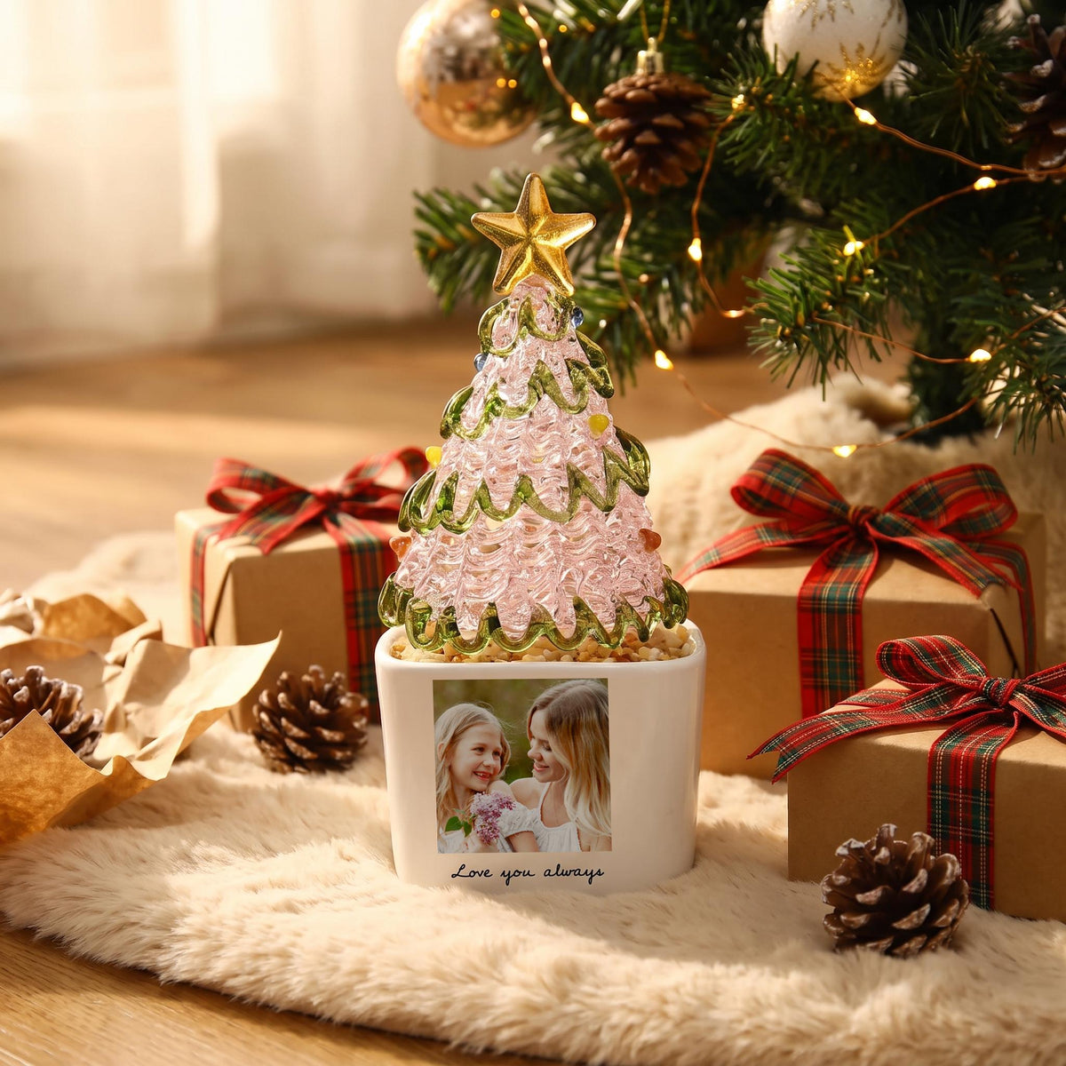 May include: A decorative glass Christmas tree with a pink textured body, green glass garland, and a gold star topper. The tree sits in a white square planter featuring a printed photograph of two people and the text Love you always.