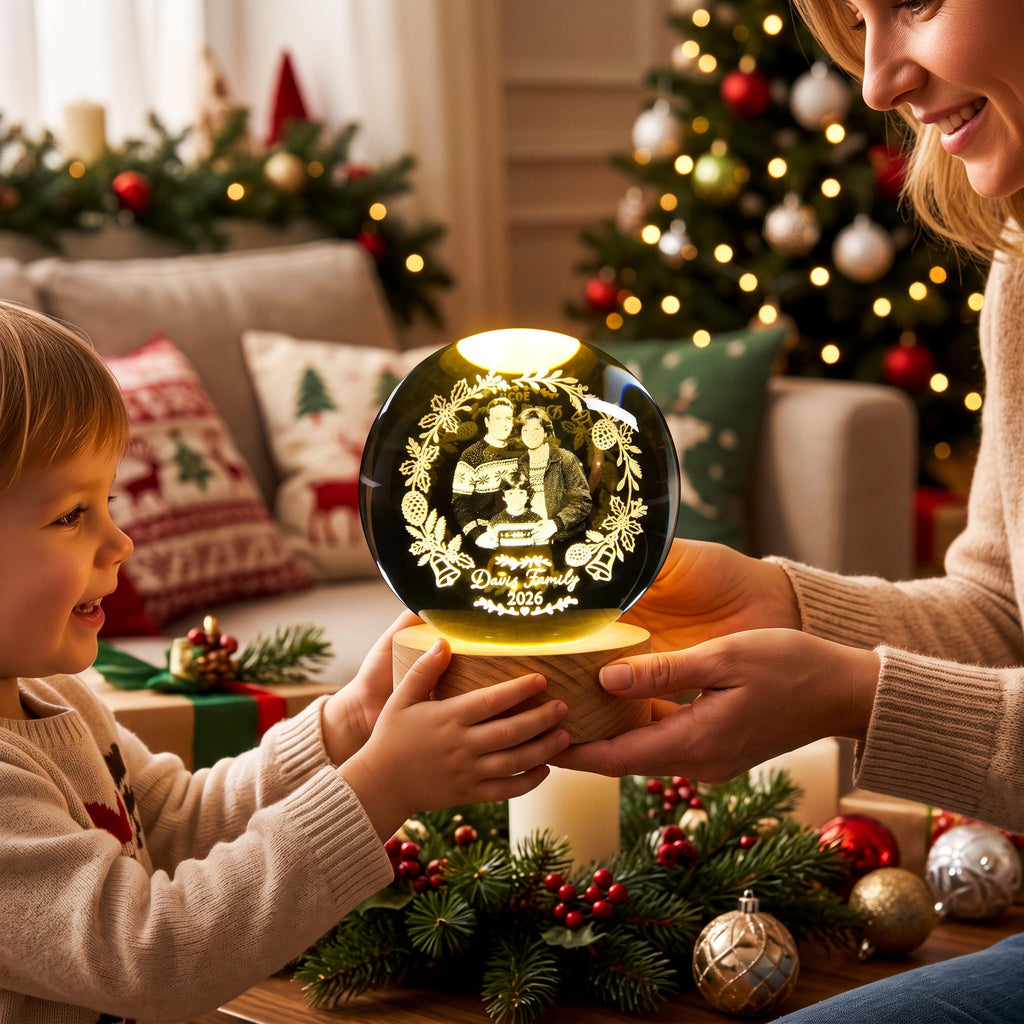 May include: A spherical glass ornament with an internal 3D laser-etched image of a family, surrounded by a wreath of pinecones and bells. The text "Davis Family 2026" is etched at the bottom. It sits on a round, light-colored wooden base.