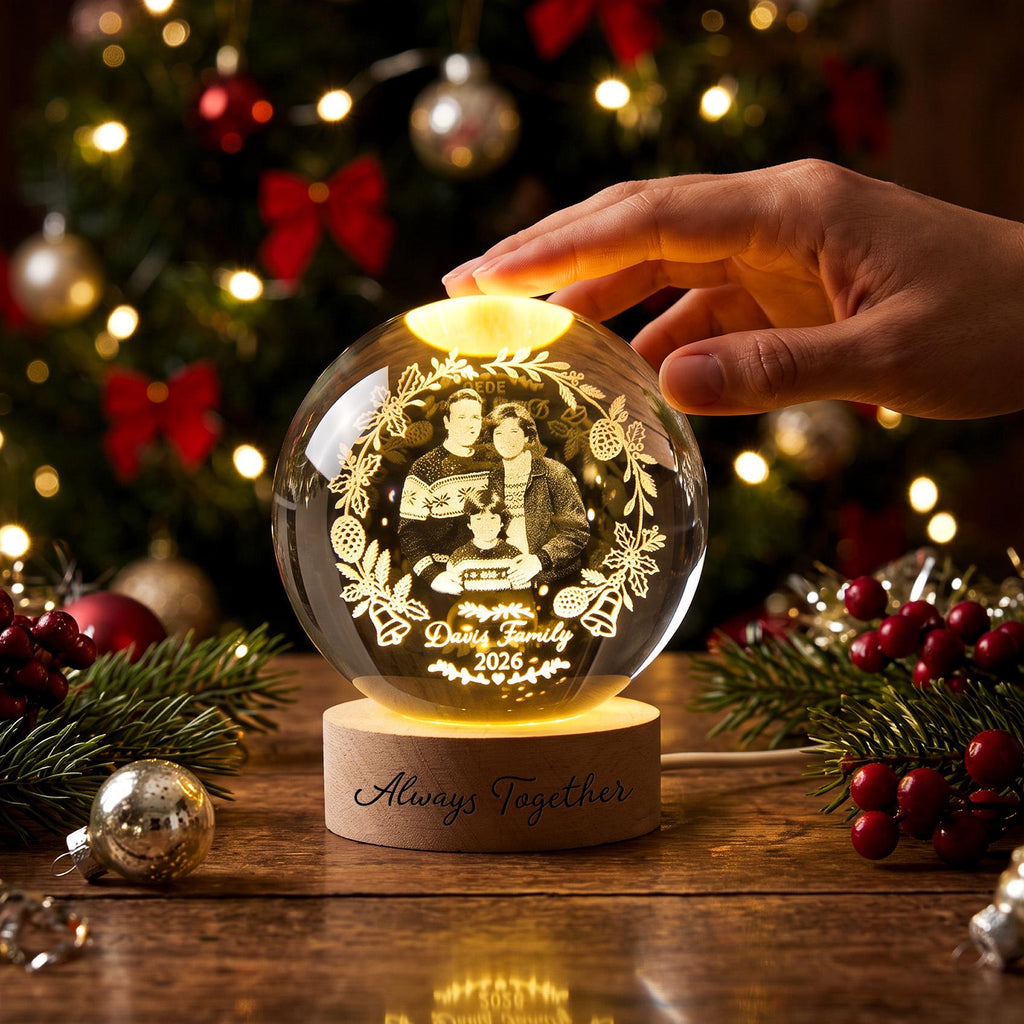 May include: A clear glass sphere featuring an internal laser-etched image of a family of three surrounded by a wreath of pinecones and bells. The sphere sits on a round wooden base engraved with the text Always Together. The text Davis Family 2026 is etched below the family portrait inside the glass.