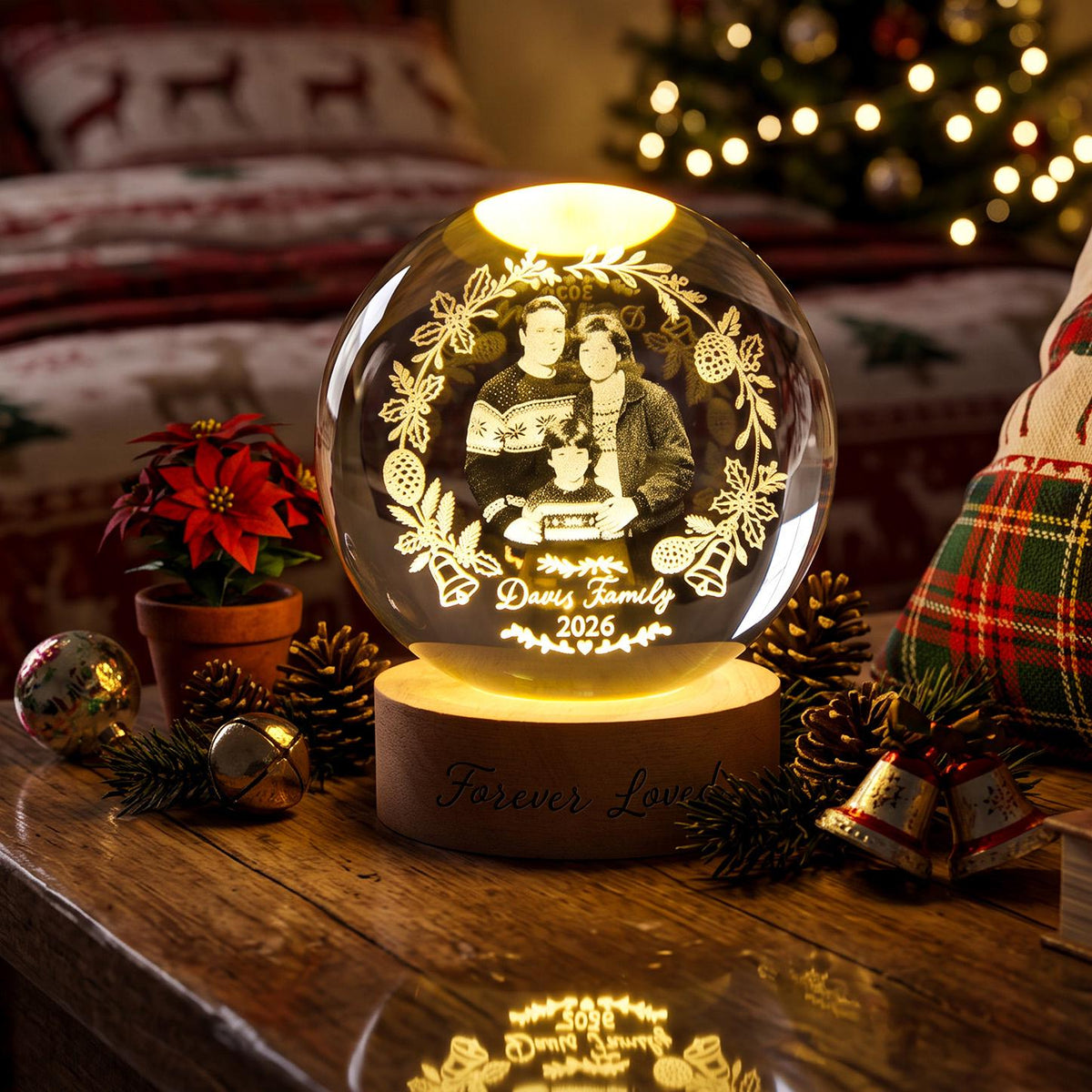 May include: A spherical glass ornament featuring an internal laser-etched image of a family of three surrounded by a wreath of holly, pinecones, and bells. The text "Davis Family 2026" is etched below. It sits on a round wooden base labeled "Forever Loved."