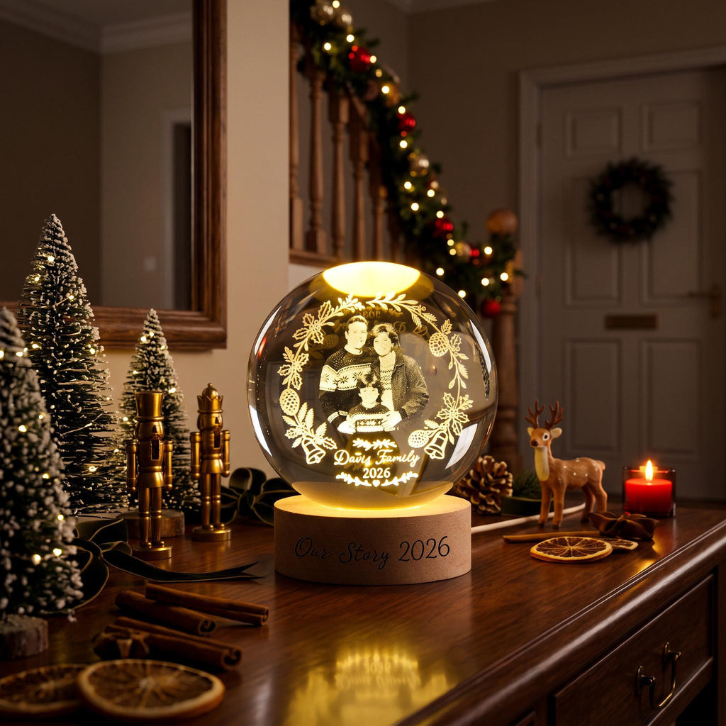 May include: A spherical glass ornament featuring an internal laser-etched image of a family within a floral wreath, inscribed with "Davis Family 2026". The glass sphere rests on a circular wooden base engraved with the text "Our Story 2026".