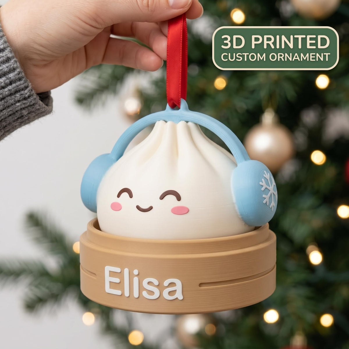 May include: A 3D printed custom ornament shaped like a white dumpling with a smiling face, wearing light blue headphones with snowflake details. The dumpling sits in a tan bamboo steamer base with the name Elisa printed in white letters.