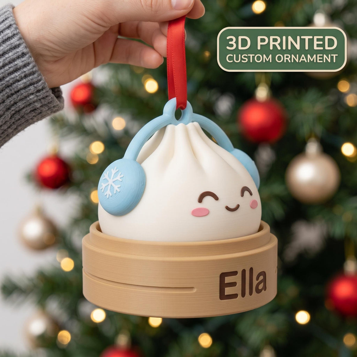 May include: A 3D printed custom ornament shaped like a white dumpling with a smiling face, wearing light blue earmuffs with snowflake details. The dumpling sits in a tan base with the name Ella printed on it, suspended by a red ribbon.
