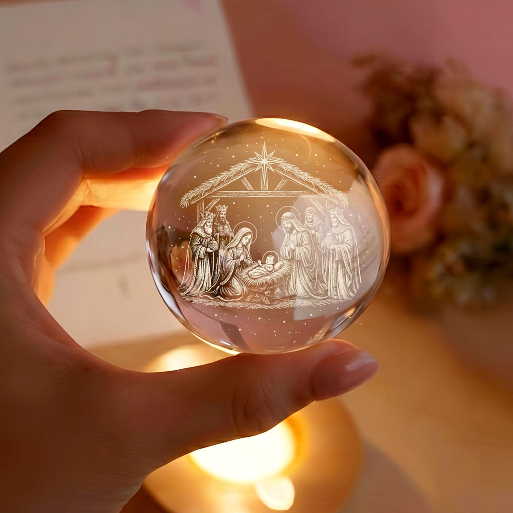 May include: A clear glass sphere featuring a laser-etched nativity scene inside. The design depicts a stable with a star above, showing figures gathered around a central manger containing a baby. The sphere is transparent with white detailing.