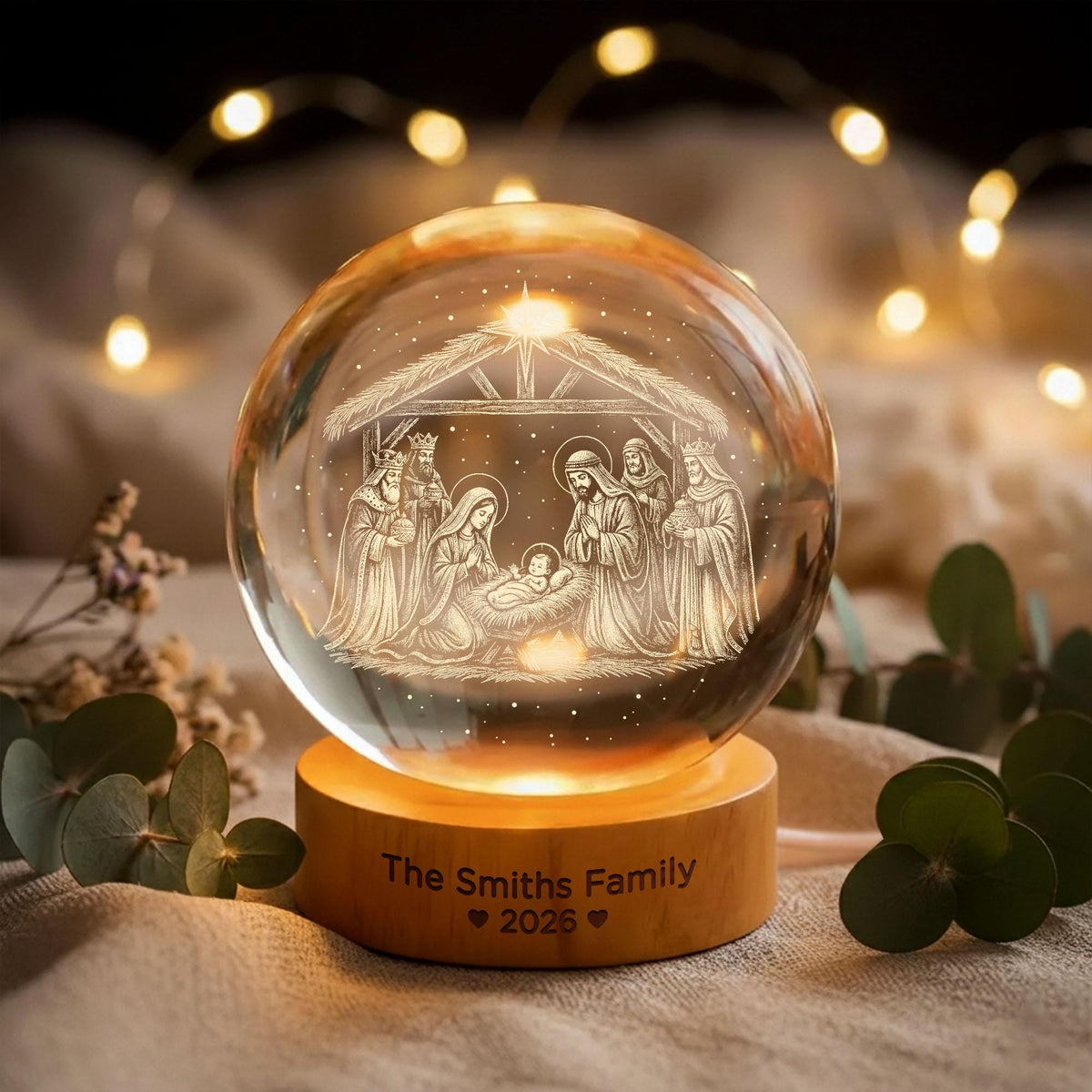 May include: A clear glass sphere featuring an internal laser-etched nativity scene with figures surrounding a manger. The sphere rests on a cylindrical wooden base engraved with the text "The Smiths Family" followed by two hearts and "2026".
