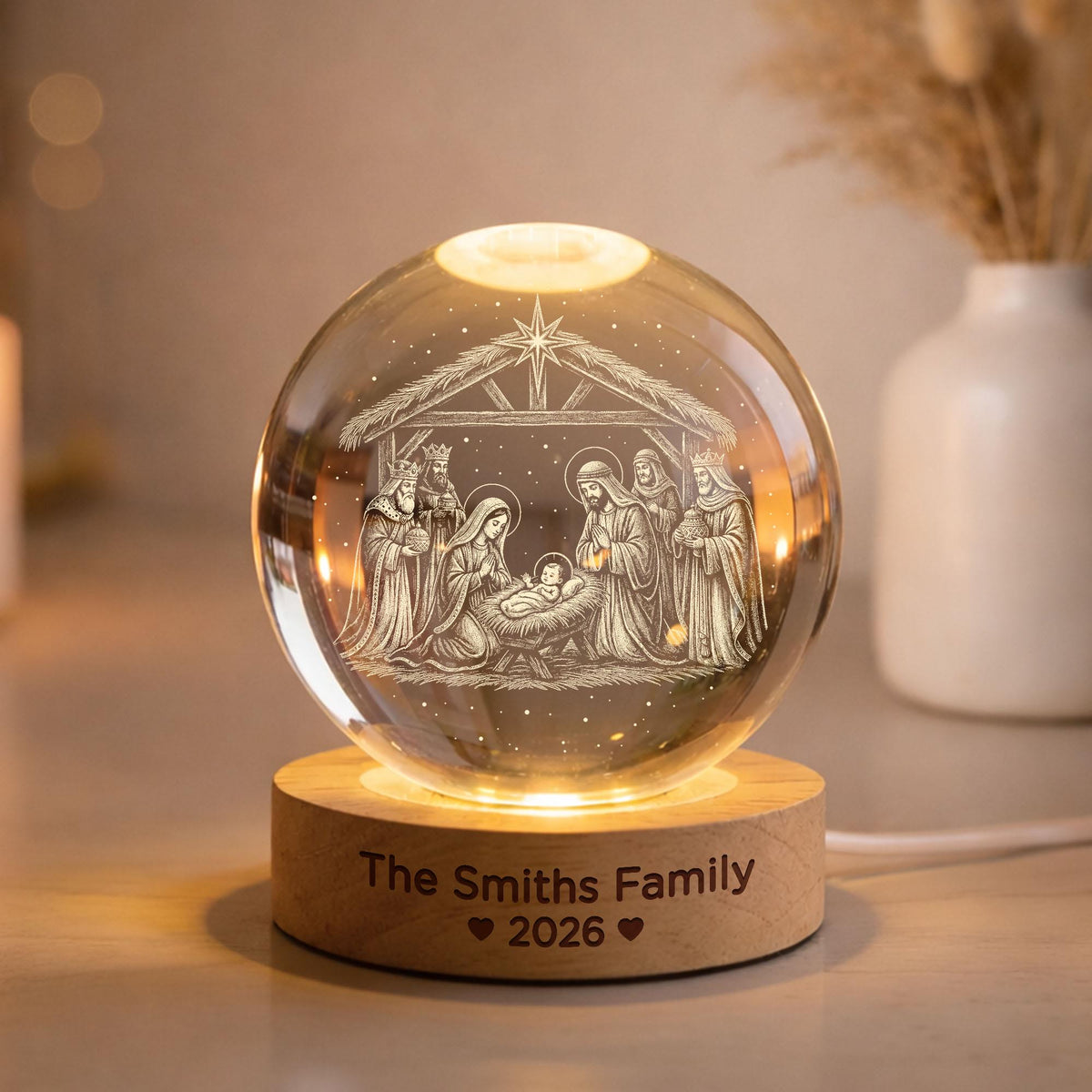 May include: A clear glass sphere featuring an internal laser-etched nativity scene with figures under a star. The sphere rests on a round wooden base engraved with the text "The Smiths Family" followed by two heart icons and the year "2026".