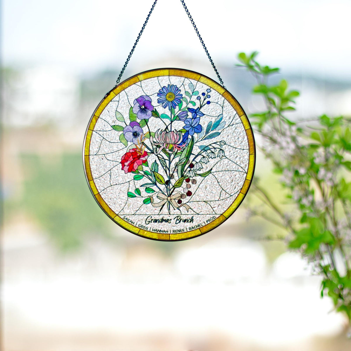 May include: A round stained glass window hanging with a floral bouquet design. The flowers include red, purple, blue, and white blooms. The text "Grandmas Brunch" is below the flowers, with names below that. The window has a gold border and is suspended by a chain.