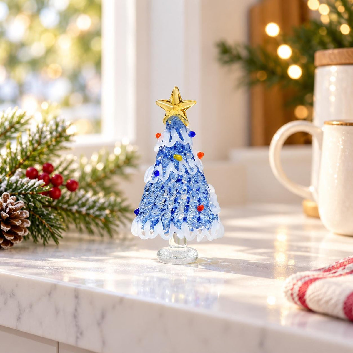 May include: A decorative glass Christmas tree figurine featuring a textured blue body with white swirling garland accents, small colorful glass bead ornaments, and a solid gold-colored star topper, all supported by a clear circular glass base.
