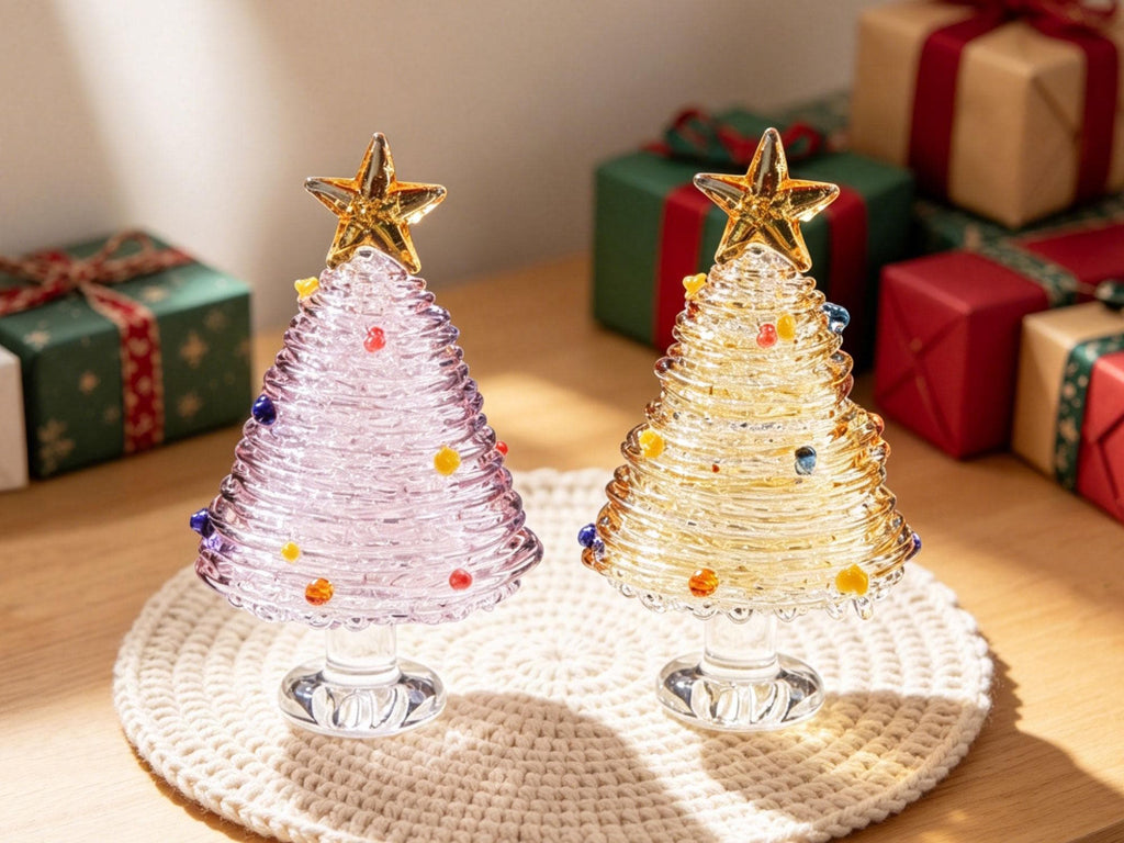 May include: Two glass Christmas tree figurines, one in light purple and one in yellow, each featuring a spiral design, small colorful glass bead ornaments, and a clear glass star topper. Both trees are mounted on a clear, circular glass base.