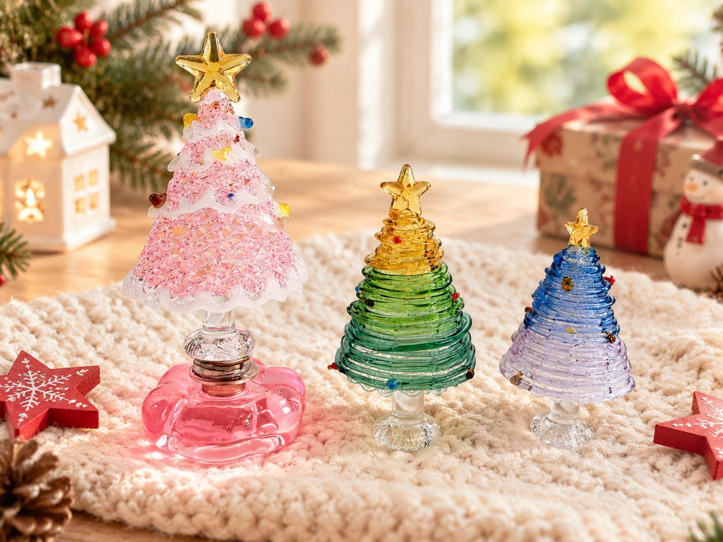 May include: Three glass Christmas tree figurines in varying sizes. The largest is pink with a textured surface, the middle is green with a coiled design, and the smallest is blue and purple. Each tree is topped with a small yellow glass star.
