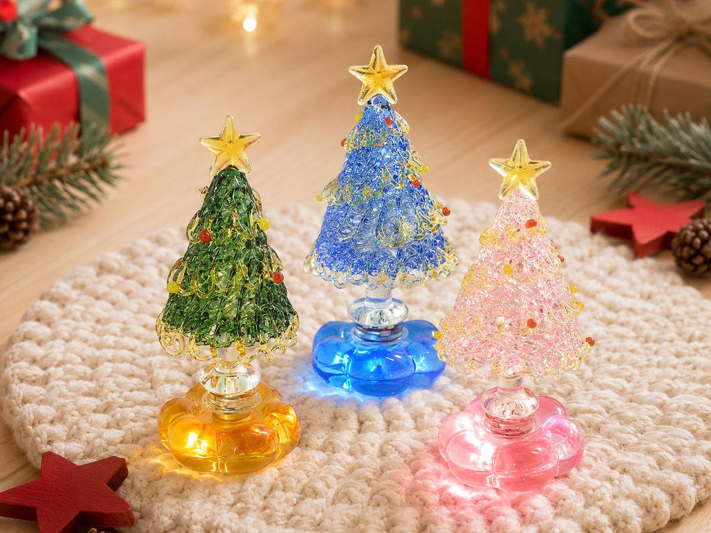 May include: Three decorative glass Christmas tree figurines with textured surfaces, each topped with a yellow star. The trees are green, blue, and pink, featuring small colorful accents and resting on translucent, flower-shaped bases.