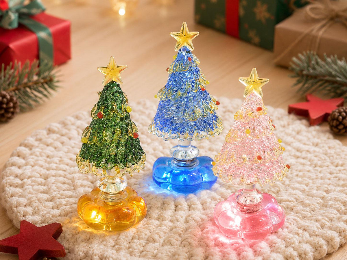 May include: Three decorative glass Christmas tree figurines with textured surfaces, each topped with a yellow star. The trees are green, blue, and pink, featuring small colorful accents and resting on translucent, flower-shaped bases.