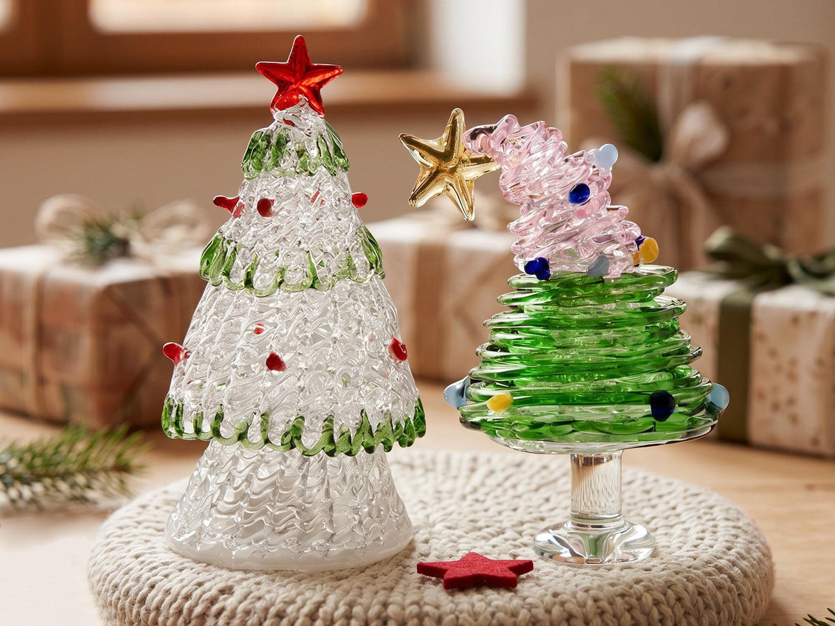 May include: Two glass Christmas tree ornaments. The left tree is clear with green trim and red accents, topped with a red star. The right tree features a green base, a pink spiraled top, a gold star, and multicolored glass dots on a pedestal.