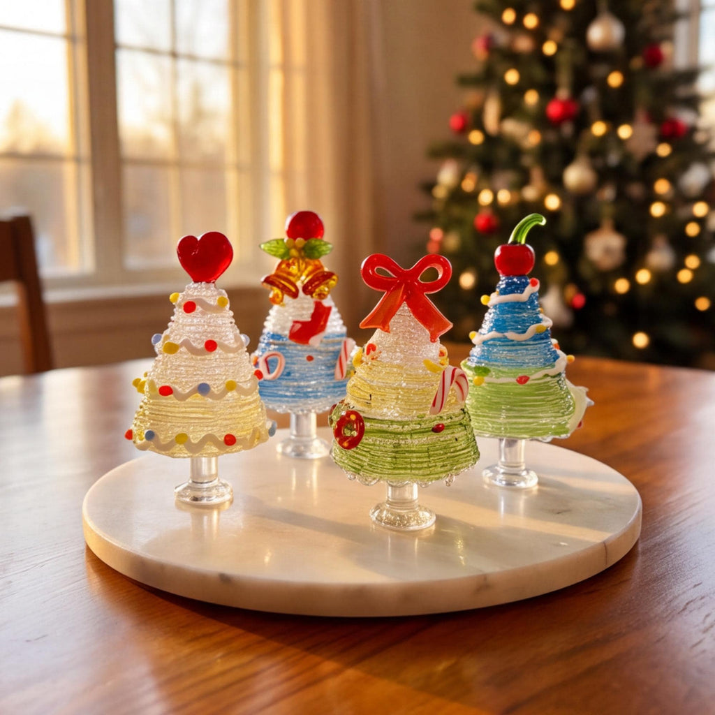 May include: Four miniature glass Christmas tree figurines on a round white marble base. Each tree features a unique topper, including a red heart, gold bells, a red bow, and a red cherry, with colorful glass accents and swirling patterns.