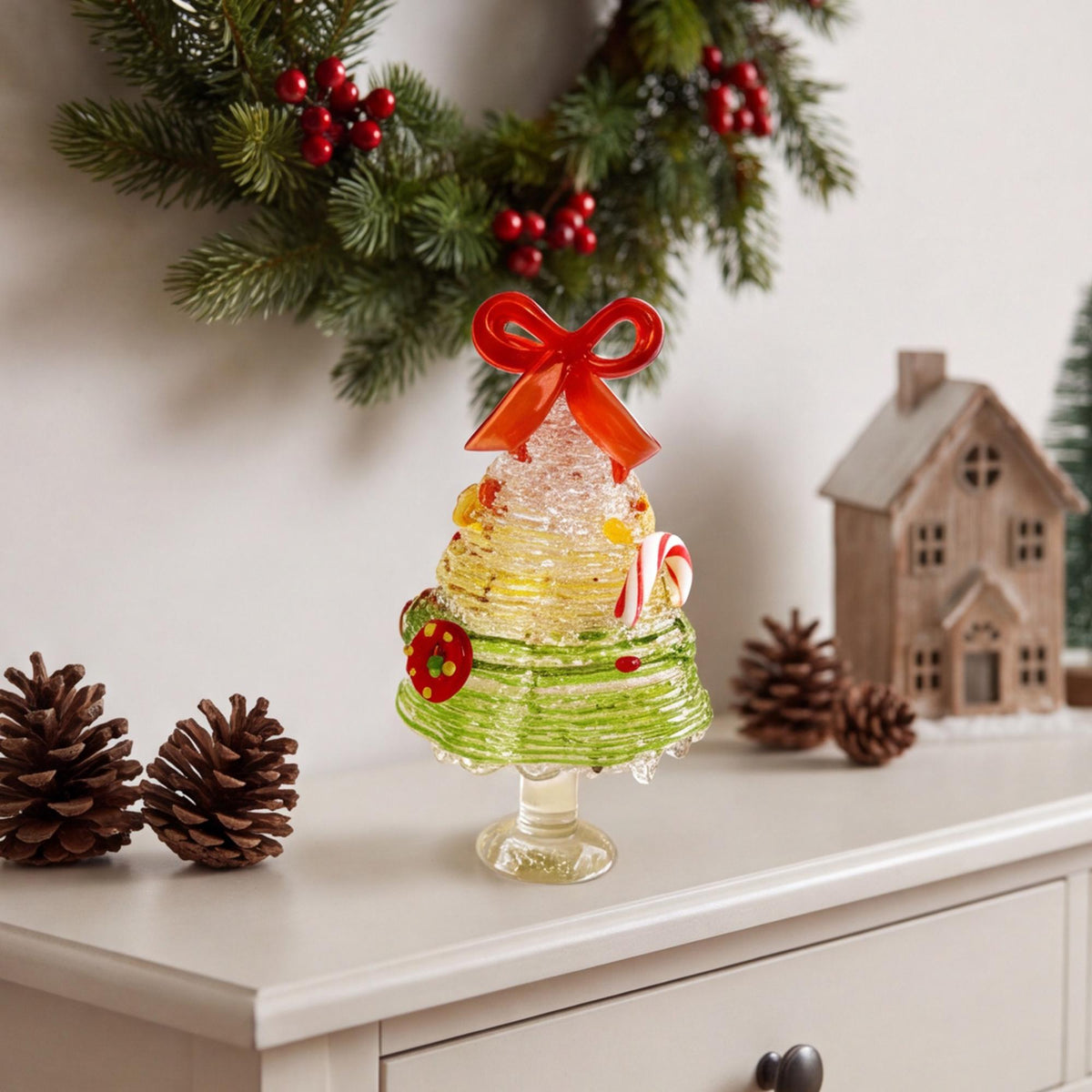 May include: A glass Christmas tree figurine featuring a red bow topper, a candy cane accent, and colorful circular ornaments. The tree has a tiered, spiraled design with a clear glass base, showcasing a gradient of white, yellow, and green colors.