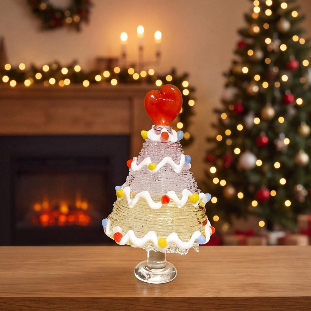 May include: A clear glass decorative tree ornament featuring a textured, tiered conical shape. The tree is adorned with white wavy glass garlands and small, colorful glass spheres, topped with a prominent red glass heart.