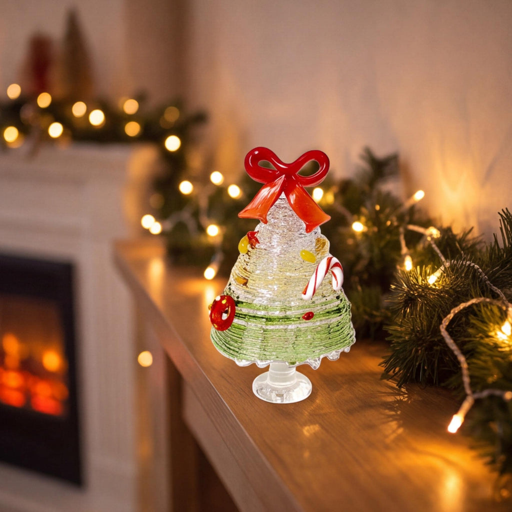 May include: A clear glass Christmas tree figurine featuring a textured, layered design with a green base, a red bow topper, and small colorful glass ornaments including a candy cane, a red ring, and yellow accents, all supported by a pedestal base.