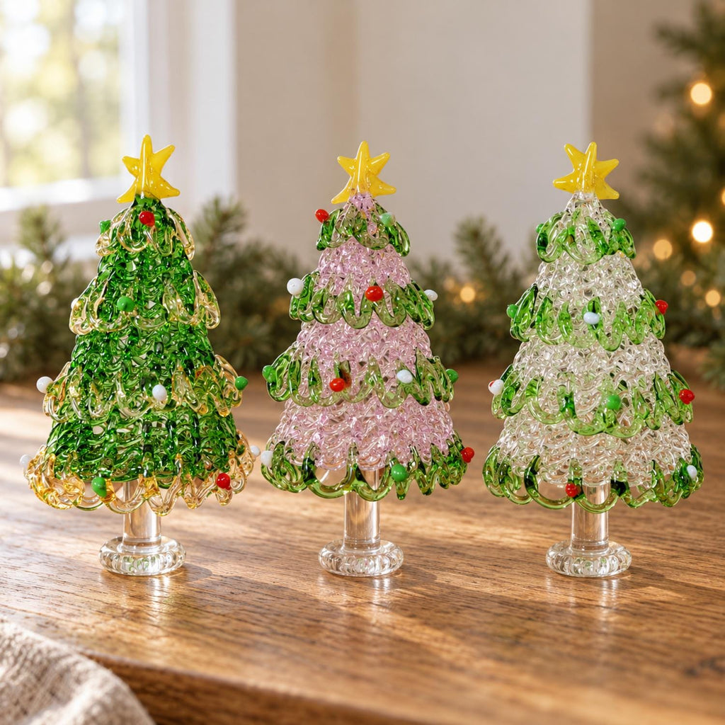 May include: Three decorative glass Christmas trees standing on clear pedestals. The left tree is green, the center is pink, and the right is clear, all featuring swirling glass branches, small colorful glass bead ornaments, and a yellow star topper.