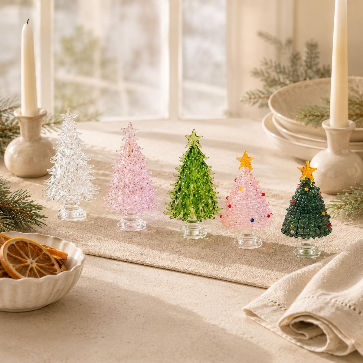 May include: Five miniature glass Christmas trees in clear, light pink, green, and dark green colors, each topped with a star. The trees feature textured, branch-like details and are arranged in a row on a table with two tall white candles.