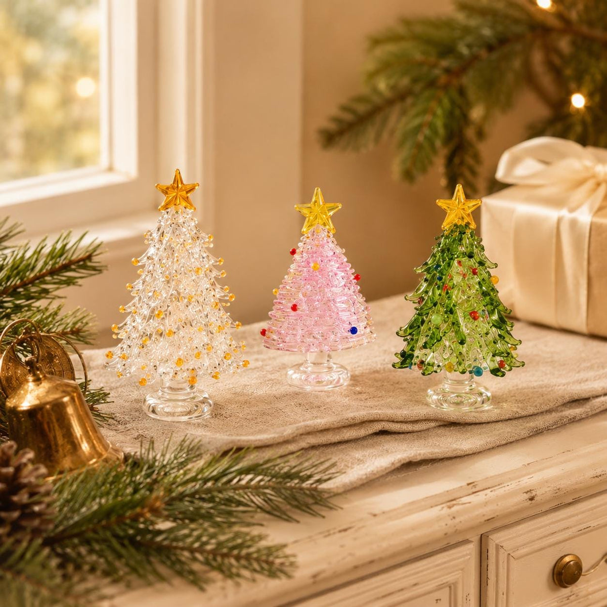 May include: Three miniature glass Christmas trees standing on a table. One tree is clear with yellow accents, one is light pink with multi-colored dots, and one is green with multi-colored dots. Each tree is topped with a yellow glass star.