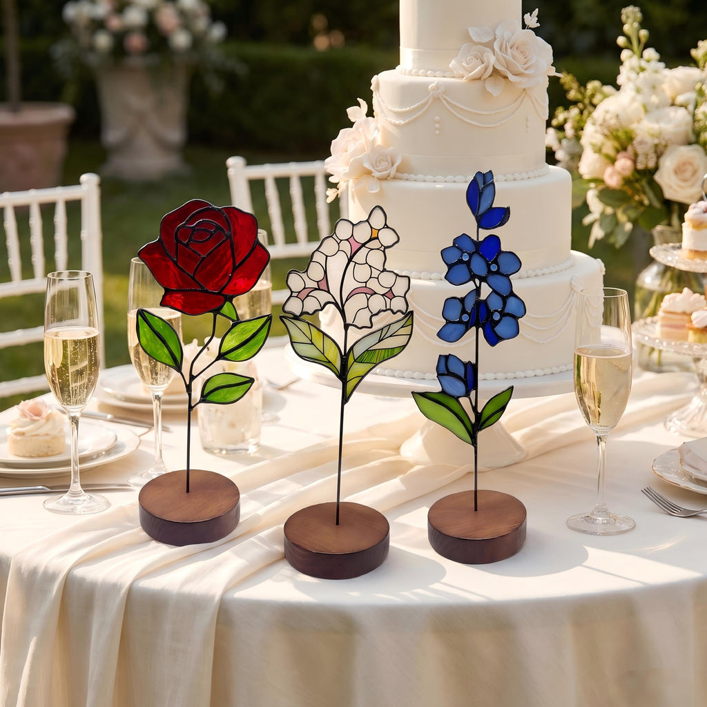 May include: Three stained glass flower sculptures on circular wooden bases. The left features a deep red rose with green leaves, the center a white hydrangea-like cluster with green leaves, and the right a tall blue delphinium-style stalk with green leaves.