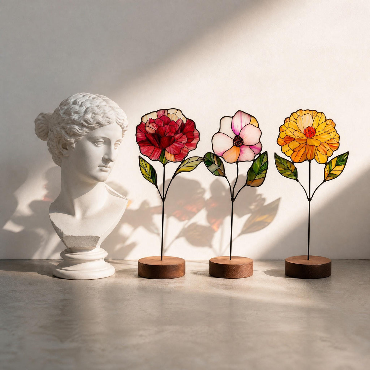 May include: A white classical bust sculpture positioned next to three stained glass flowers on thin metal stems with round wooden bases. The flowers feature red, pink, and yellow petals with green leaves, all outlined in dark metal solder lines.